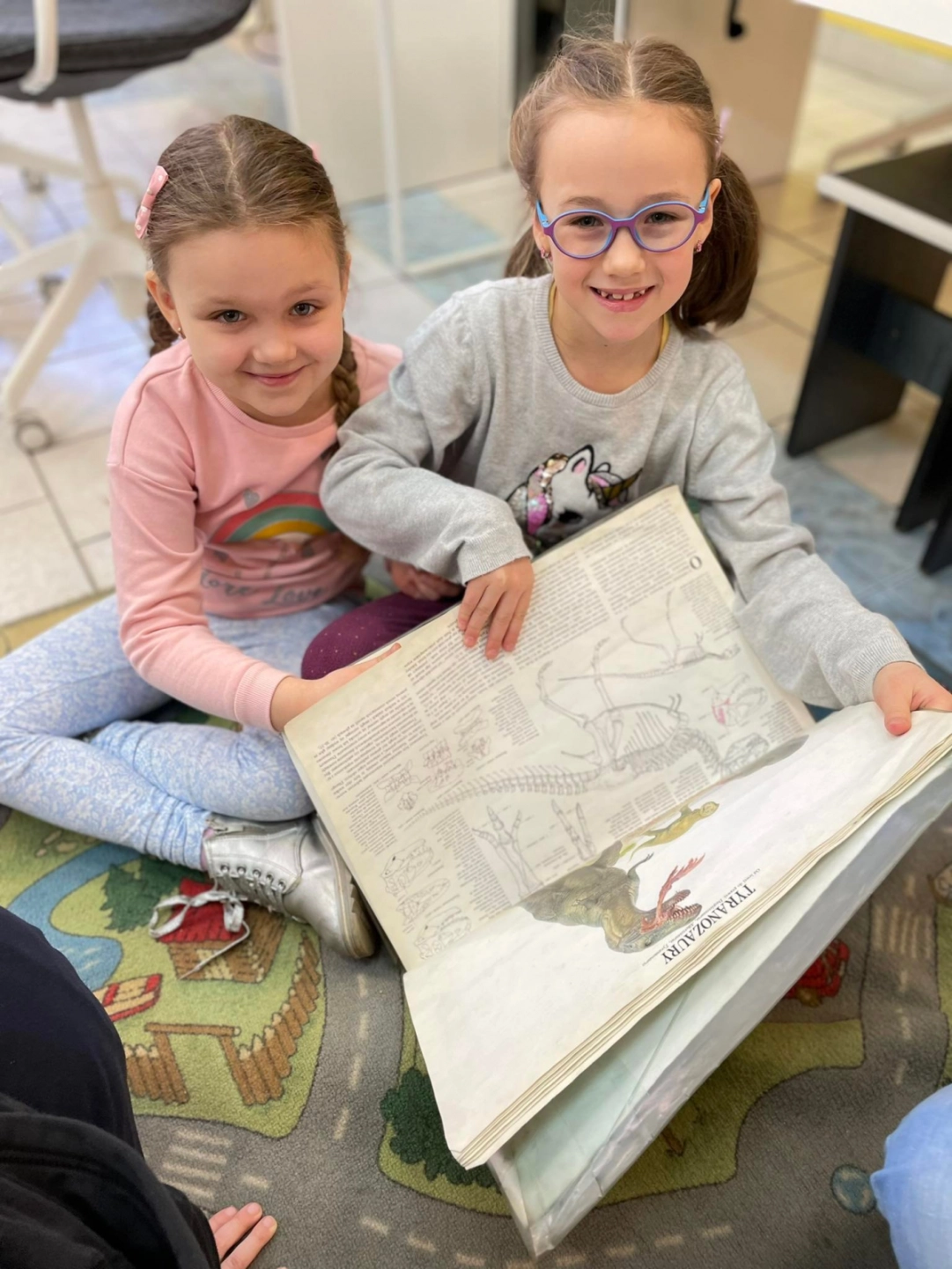 Dwie dziewczynki siedzą na dywanie i pokazują książkę o dinozaurach - powiększenie