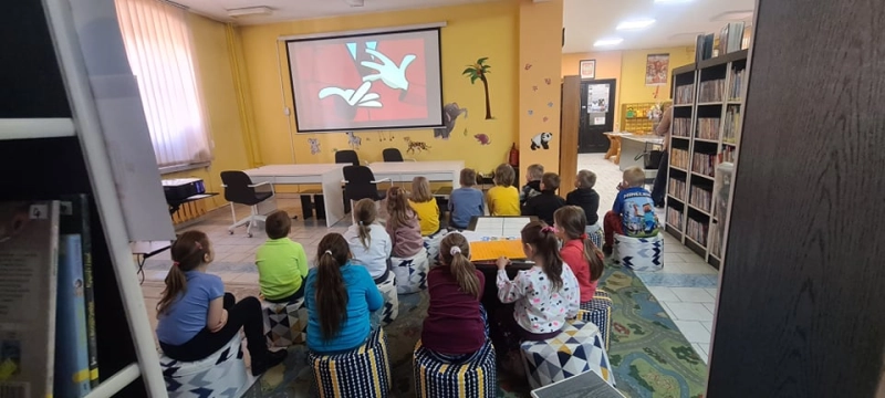 Dzieci siedzą na pufach w Oddziale dla Dzieci i oglądają prezentację multimedialną na ekranie - powiększenie