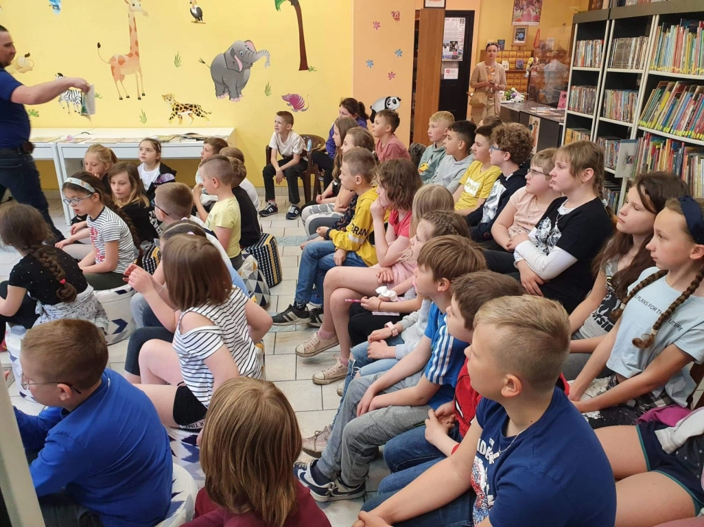 Uczniowie siedzący w Oddziale dla Dzieci słuchają Norberta Kościeszę, trzymającego w ręku książkę - powiększenie