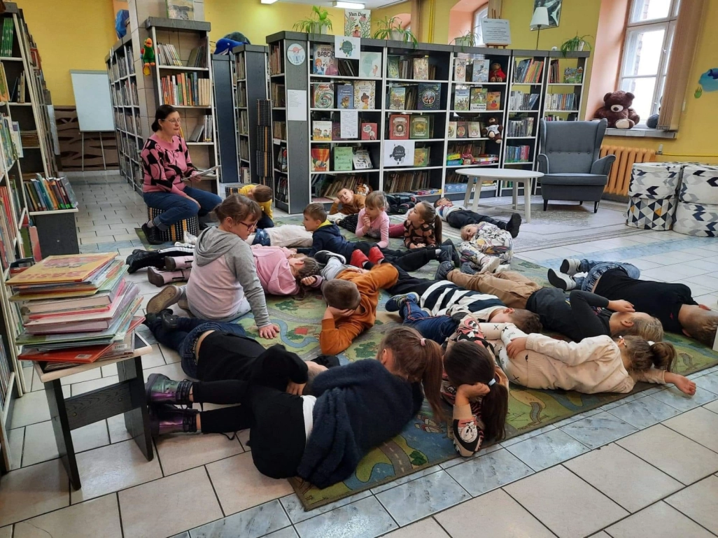 Uczniowie leżą na dywanie. Przed nimi siedzi na taborecie prowadząca zajęcia - powiększenie