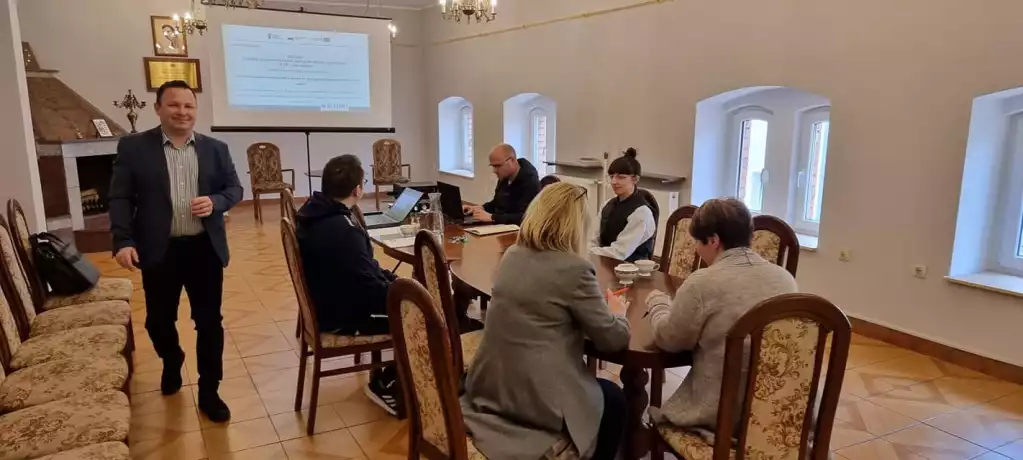 Prowadzący szkolenie Witold Gajda stoi obok siedzących przy stole pracownikach biblioteki. - powiększenie