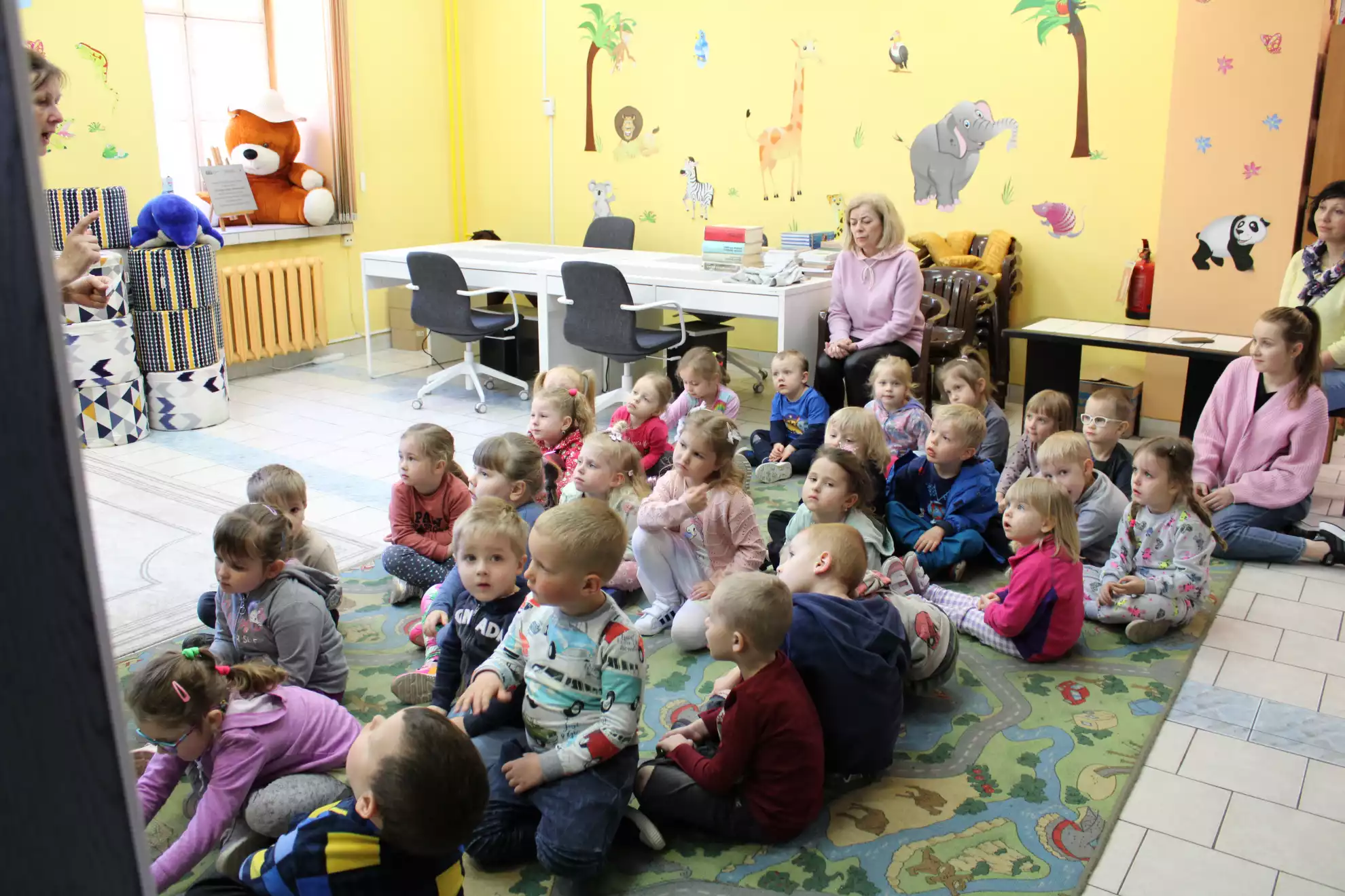 Dzieci siedzą na dywanie i słuchają pani bibliotekarki - powiększenie
