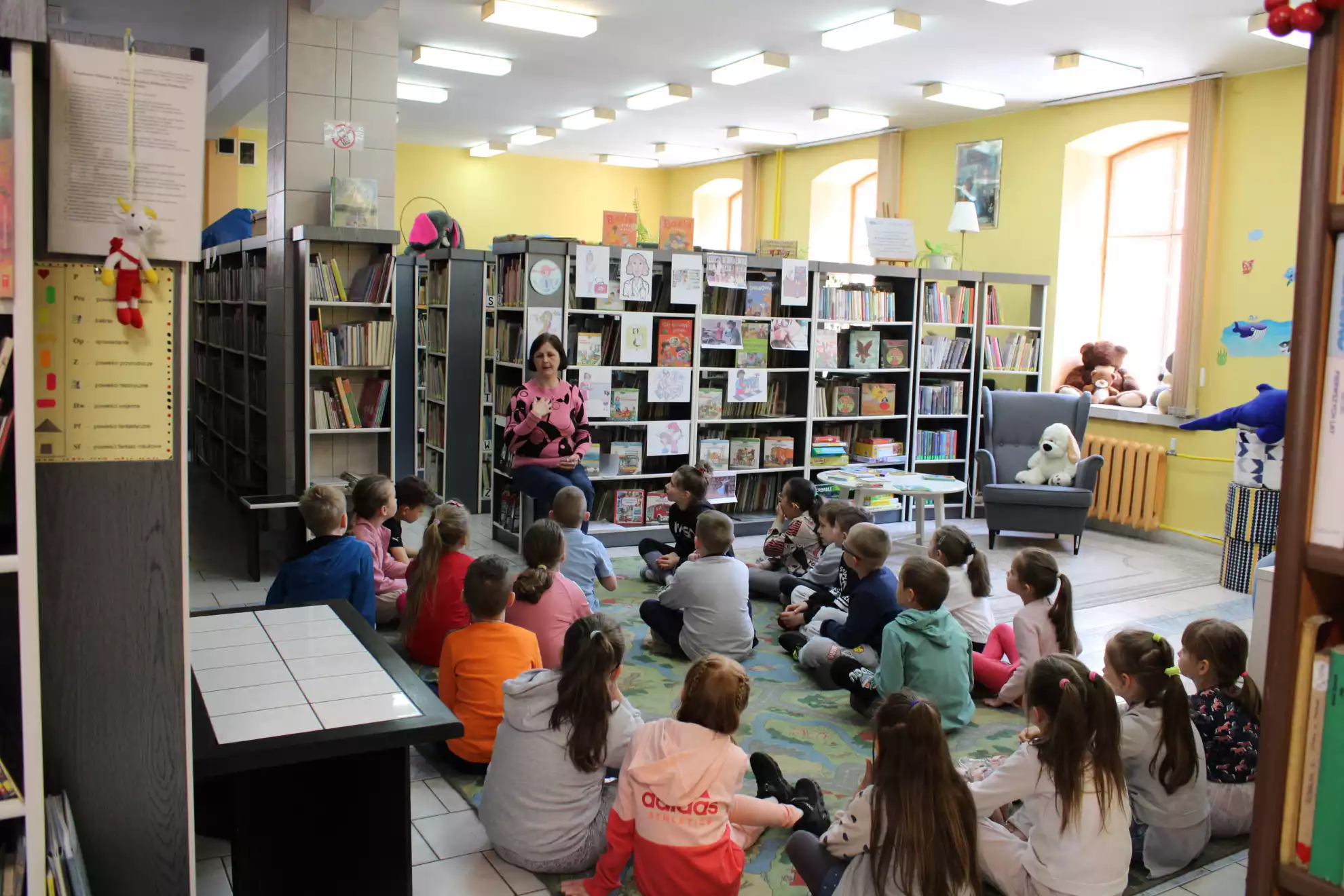 Dzieci siedzą na dywanie i słuchają pani bibliotekarki - powiększenie