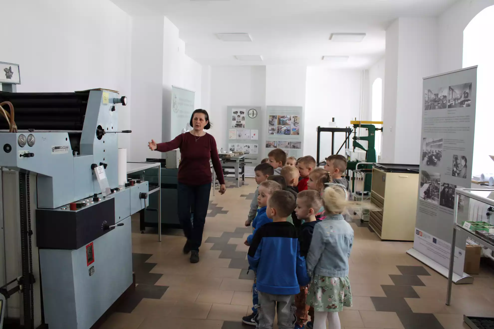 Dzieci wraz z panią bibliotekarką oglądają maszyny drukarskie  - powiększenie