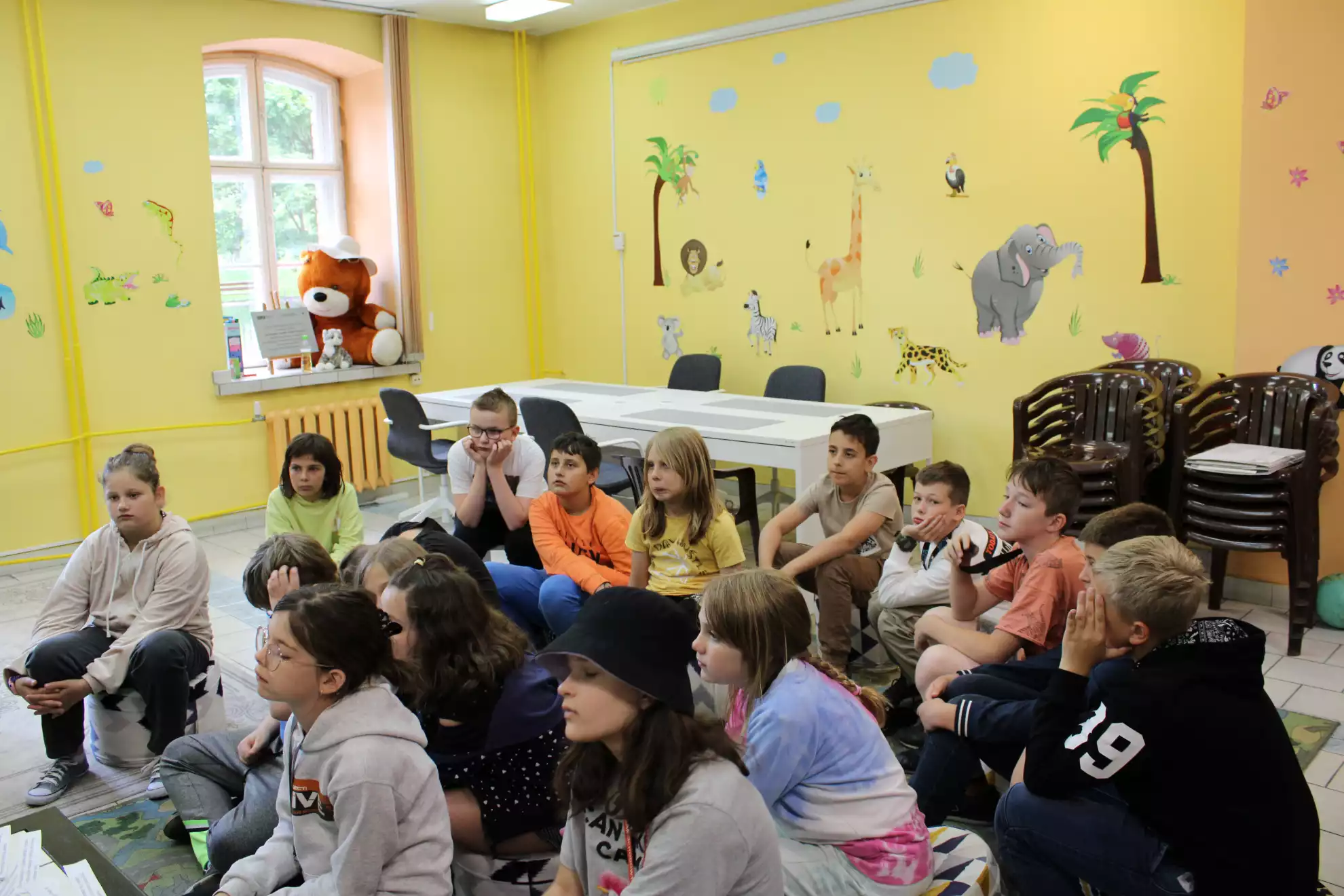 Grupa uczniów siedzi na pufach w Oddziale dla Dzieci, słuchając prowadzącej warsztaty. - powiększenie