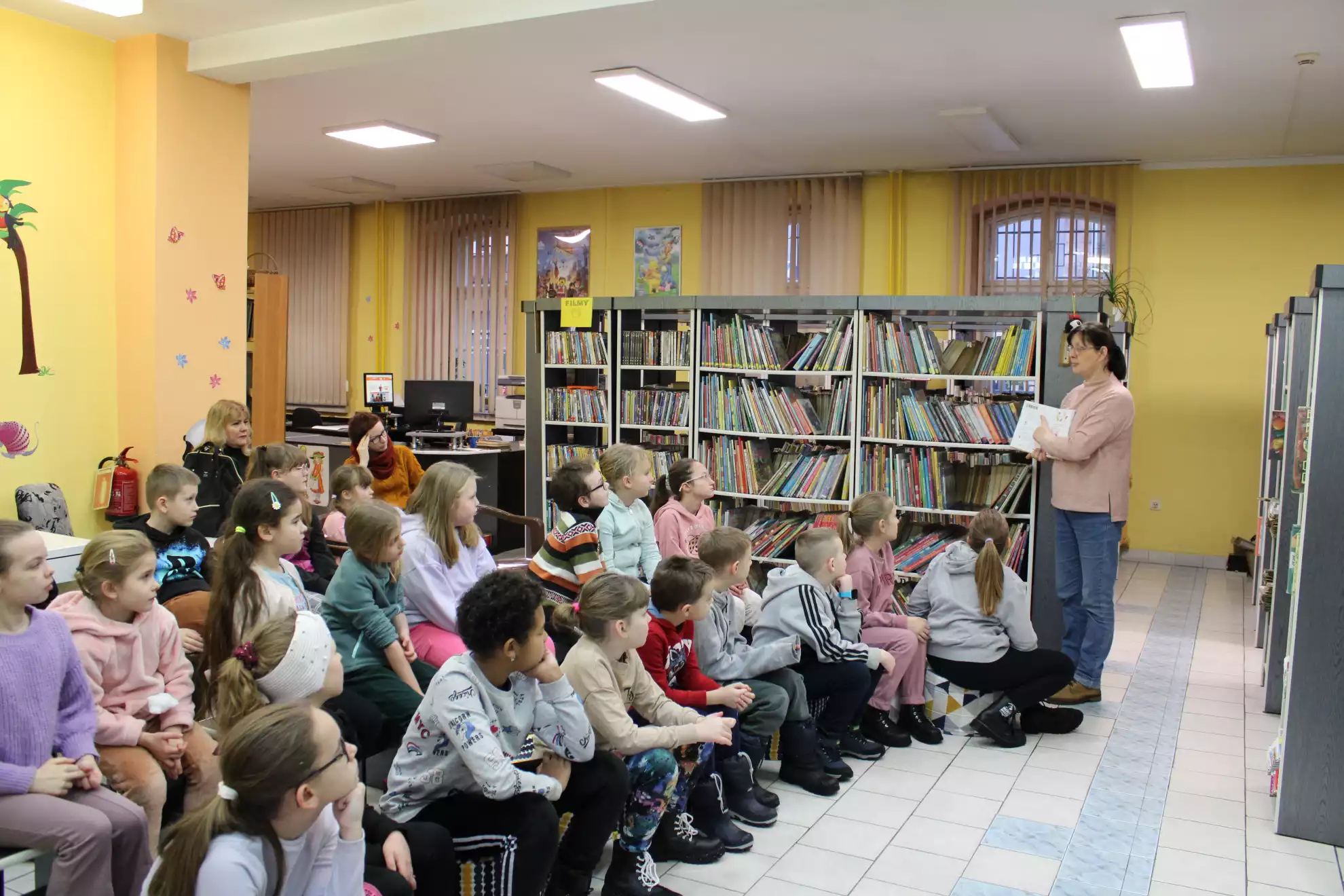 Dzieci siedzą w Oddziale dla Dzieci i słuchają jak wygląda praca w bibliotece. - powiększenie