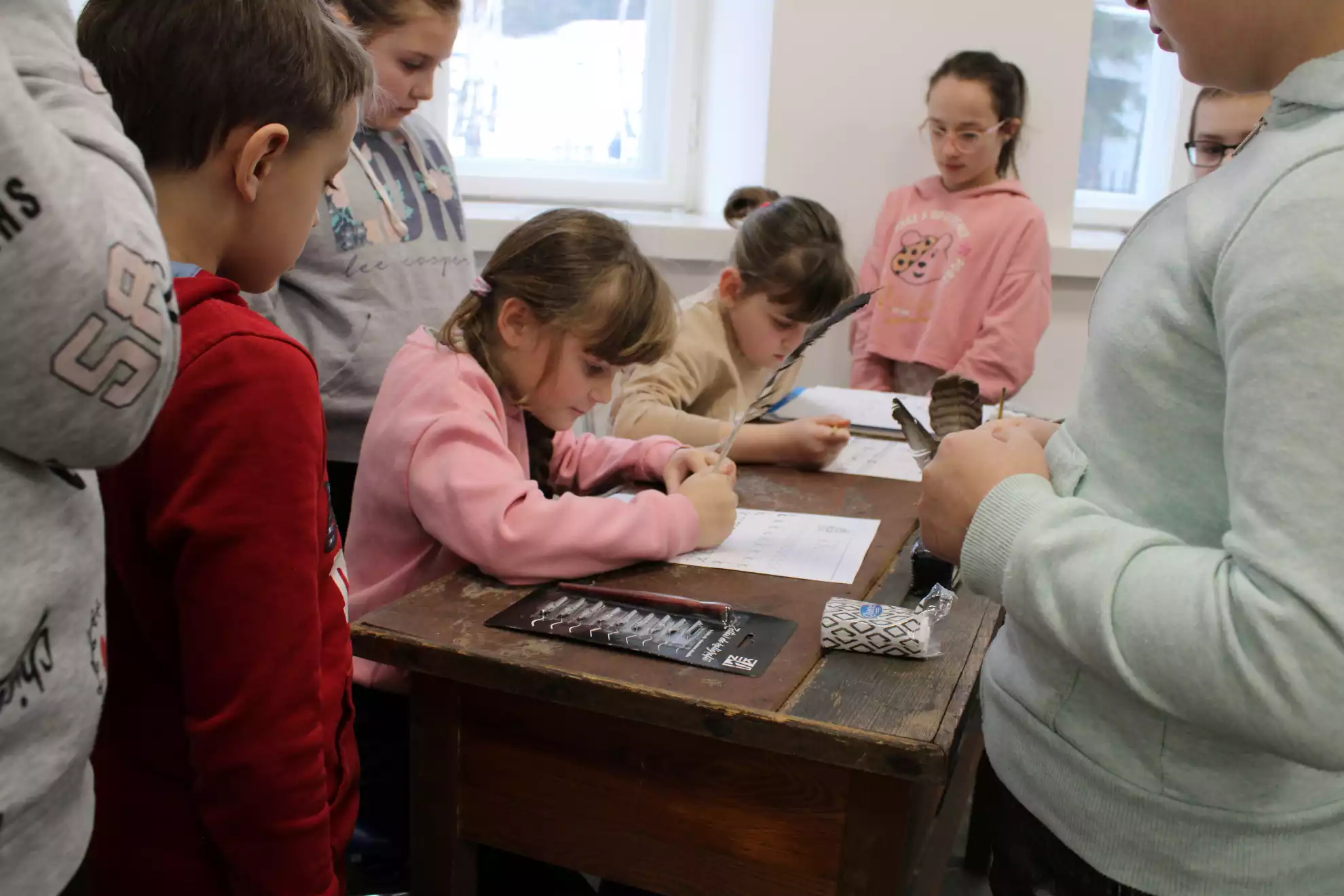 Dzieci siedzą przy starym biurku w Pracowni Poligrafii i ćwiczą sztukę kaligrafii.  - powiększenie