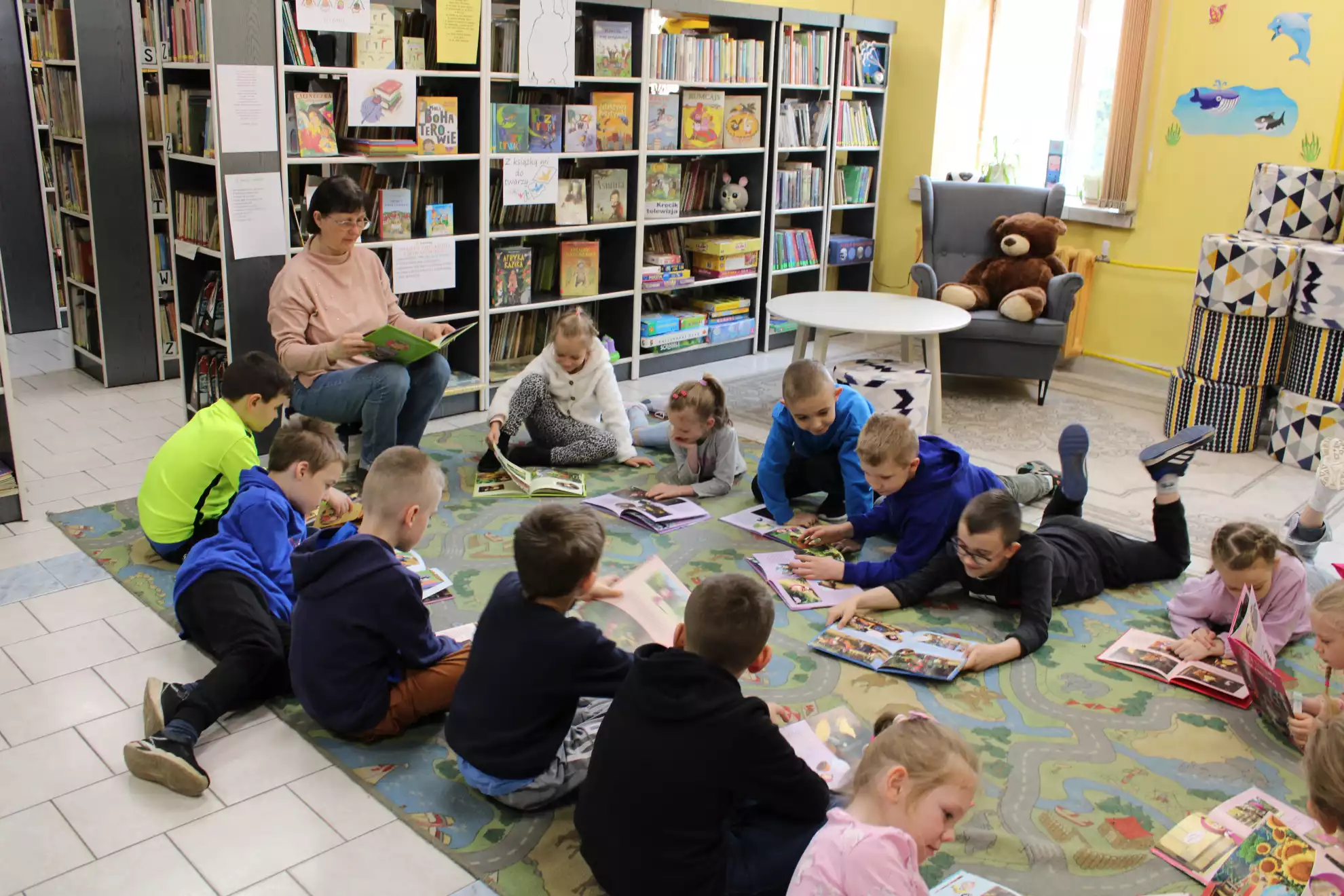 Dzieci siedzą na dywanie z książkami i słuchają pani bibliotekarki. - powiększenie