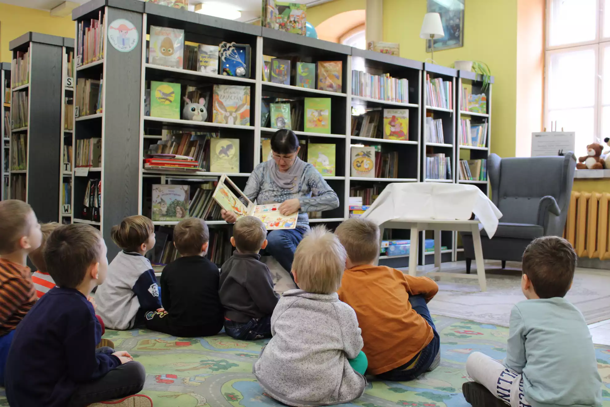 Dzieci siedzą na dywanie i oglądają książkę, która pokazuje pani bibliotekarka.  - powiększenie