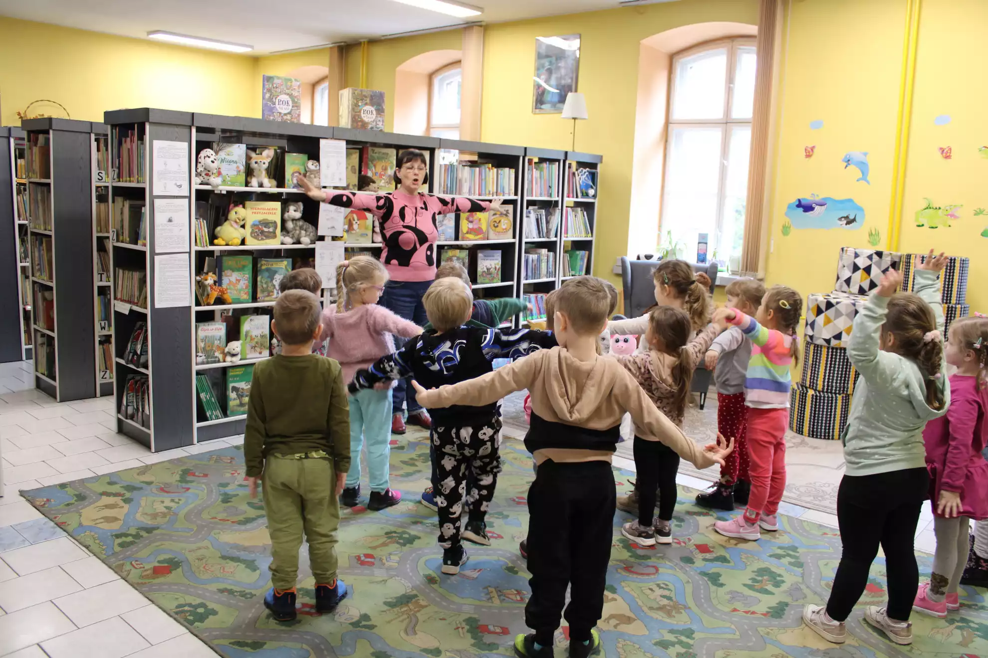 Dzieci stoją na dywanie i powtarzają ruchy wykonywane przez panią bibliotekarkę.  - powiększenie