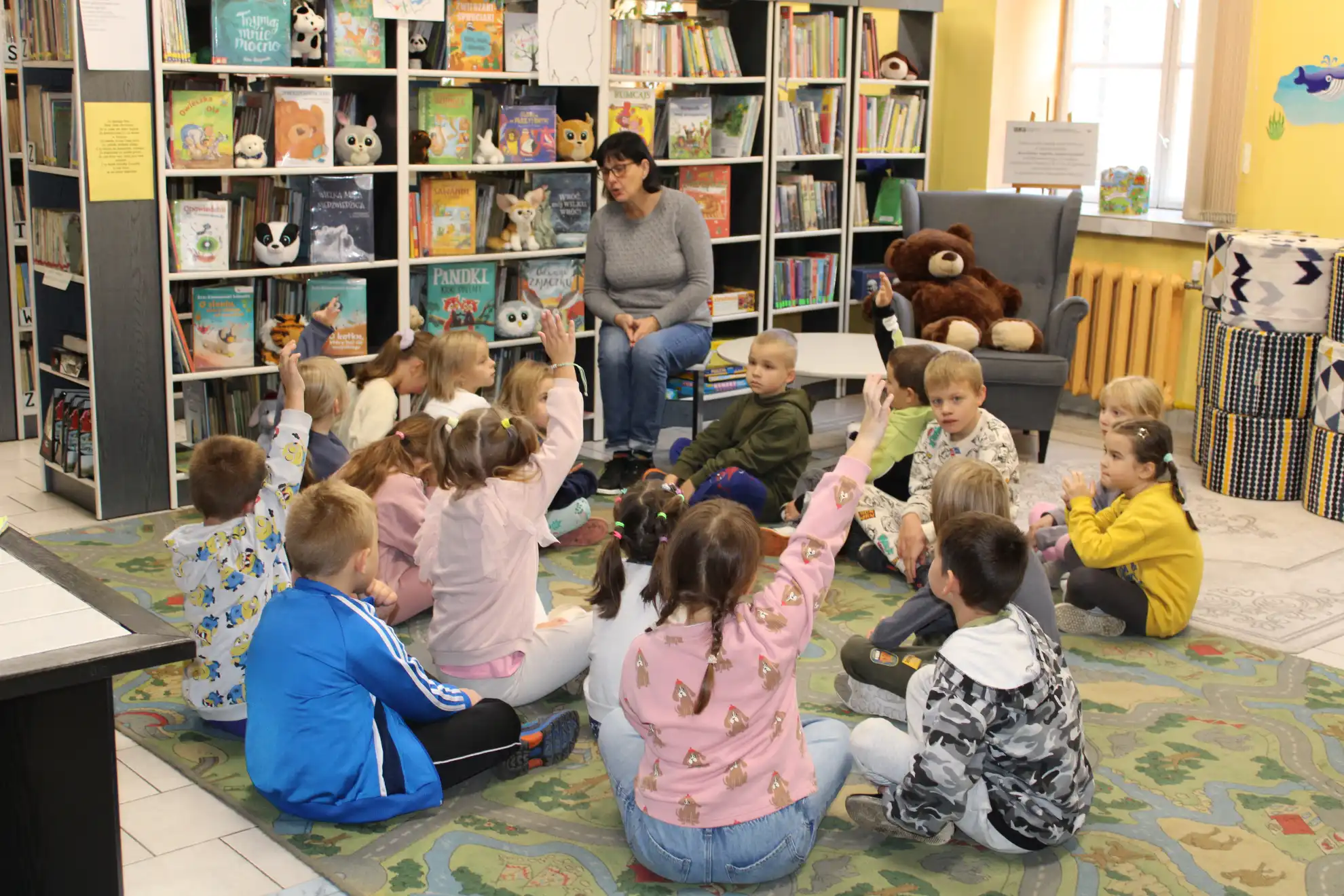 Dzieci siedzą na dywanie i słuchają pani bibliotekarki.  - powiększenie