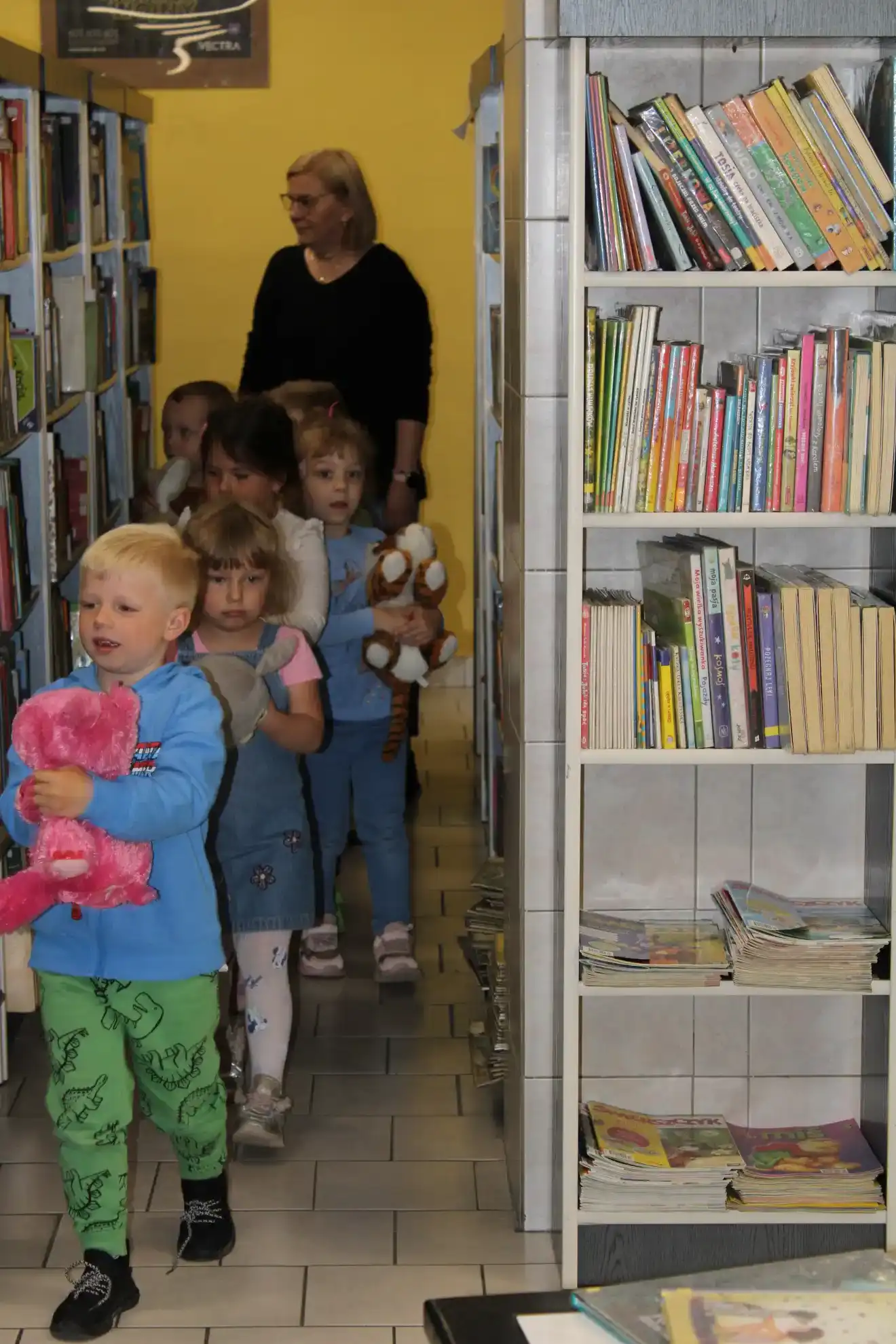 Dzieci z pluszakami w ręku spacerują po bibliotece.  - powiększenie