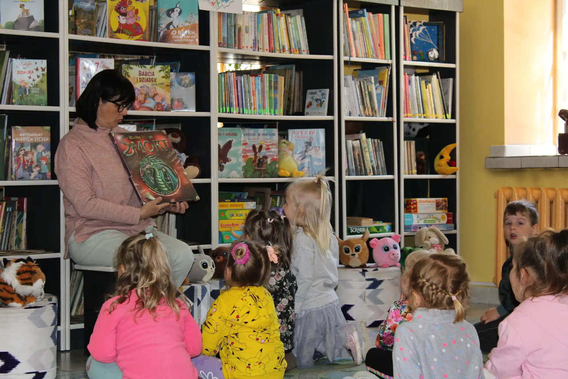 Dzieci siedzą na dywanie i oglądają książkę pokazywaną przez panią bibliotekarkę.  - powiększenie