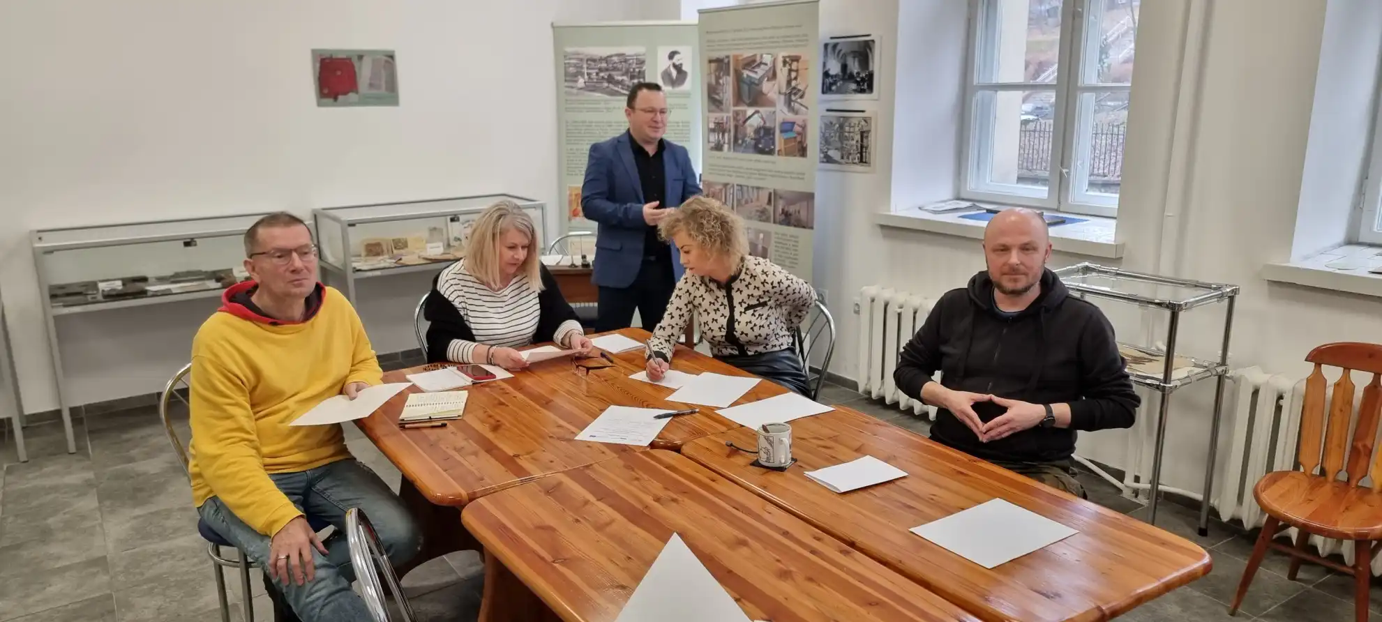 Bibliotekarze siedzą przy stole w trakcie szkolenia. Na drugim planie stoi prowadzący. - powiększenie