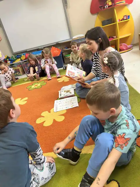 Pani bibliotekarka siedzi z dziećmi na dywanie i czyta bajkę. 