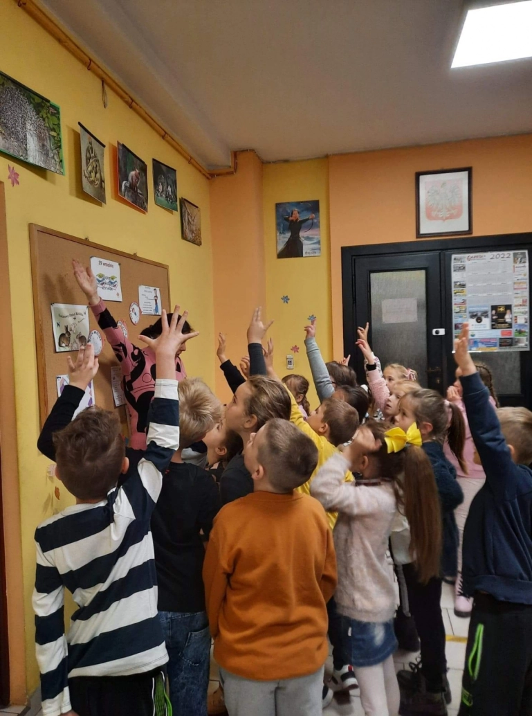 Uczniowie stojący przed wystawą swoich prac, zgłaszają się trzymając rękę w górze