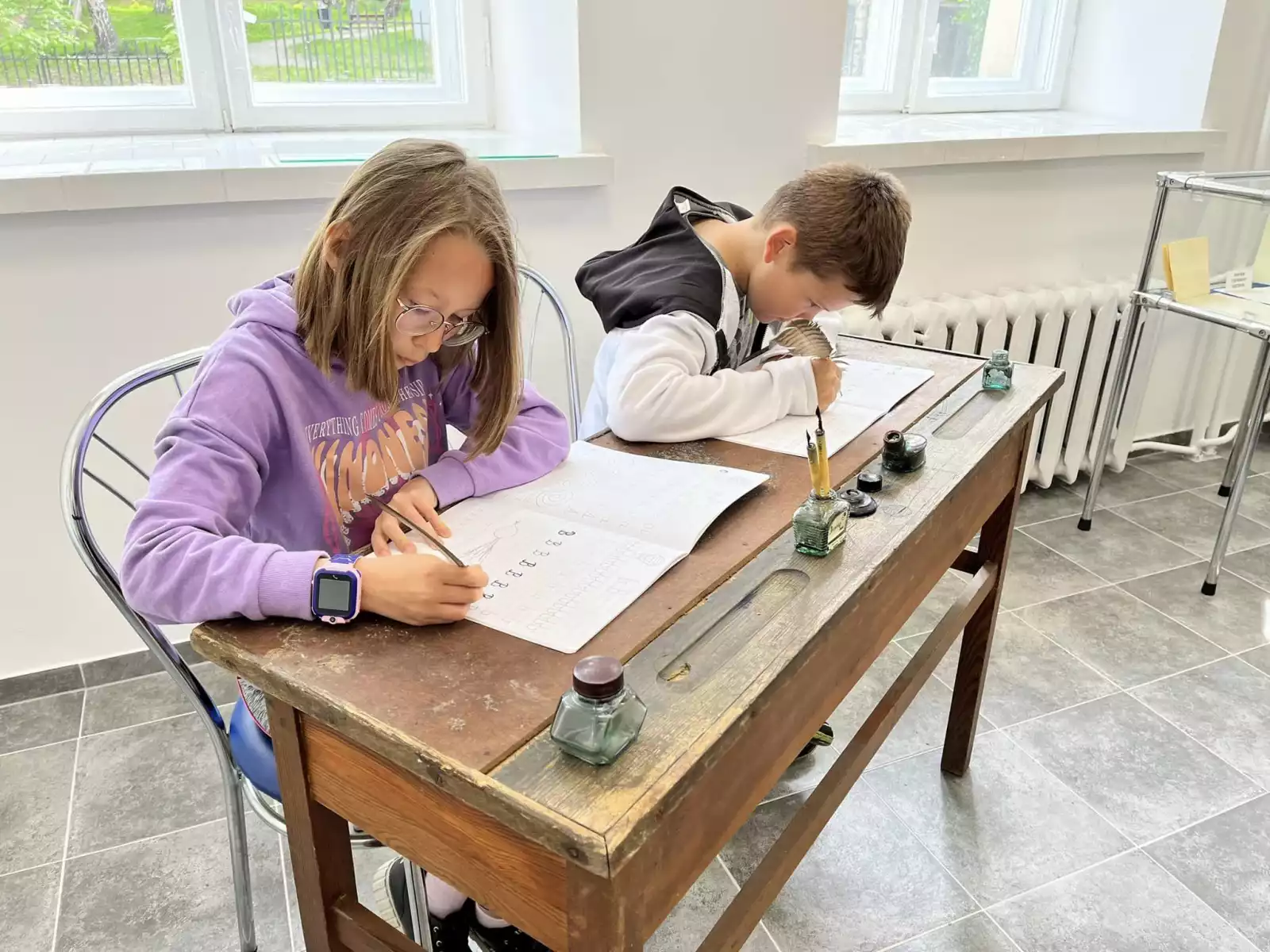 Dwójka dzieci trenuje kaligrafię, siedząc przy zabytkowej ławce szkolnej.