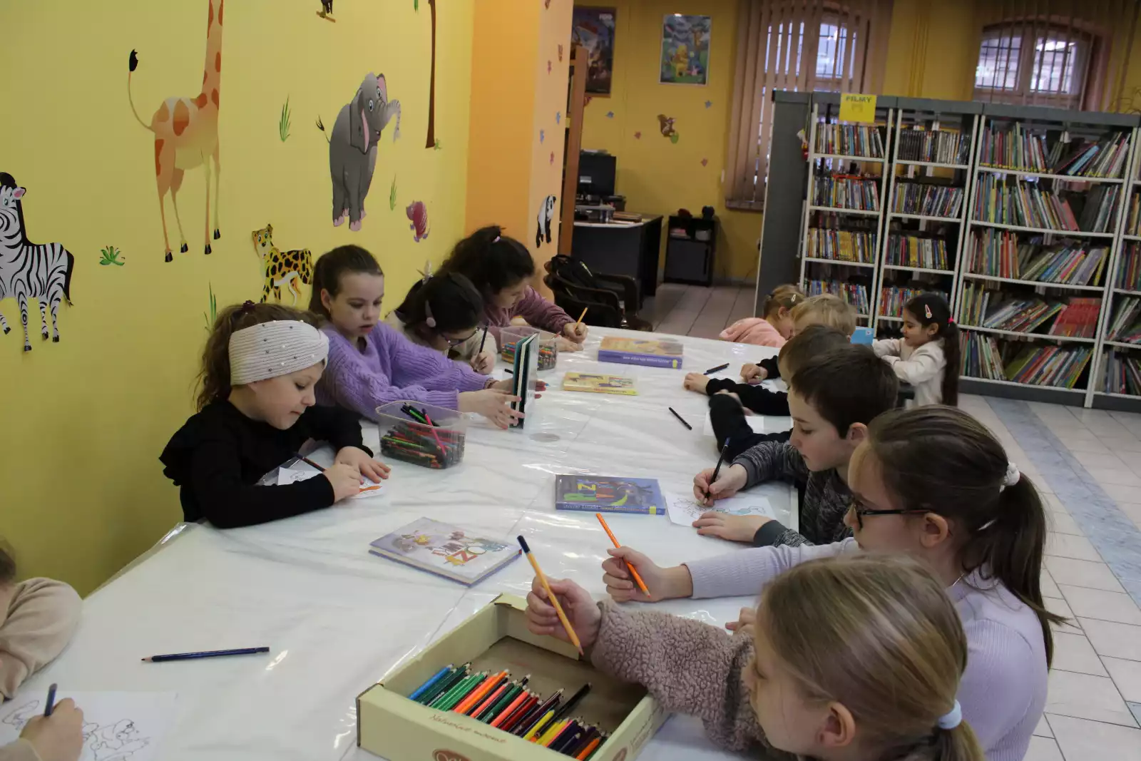 Dzieci siedzą przy stolikach w Oddziale dla Dzieci i kolorują ilustracje do książki. 