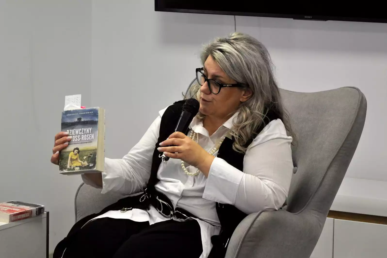 Agnieszka Dobkiewicz siedzi w fotelu. W jednej ręce trzyma swoją książkę, a w drugiej mikrofon.