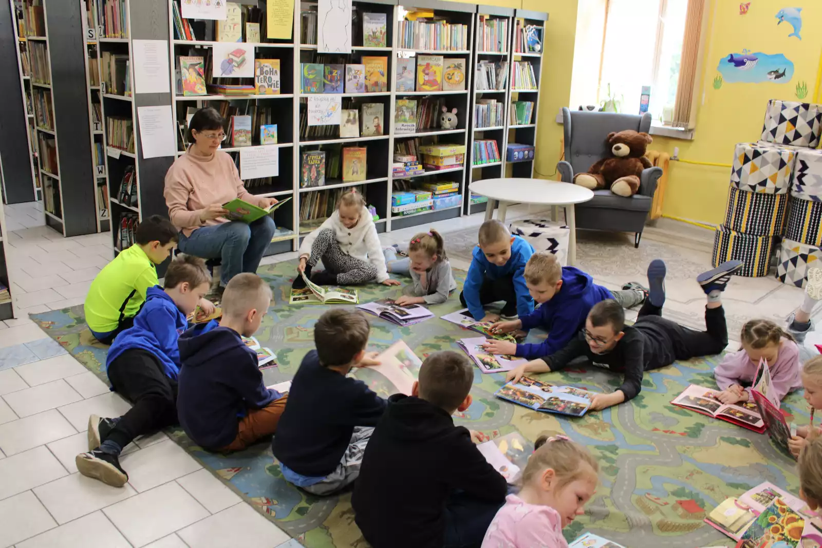 Dzieci siedzą na dywanie z książkami i słuchają pani bibliotekarki.