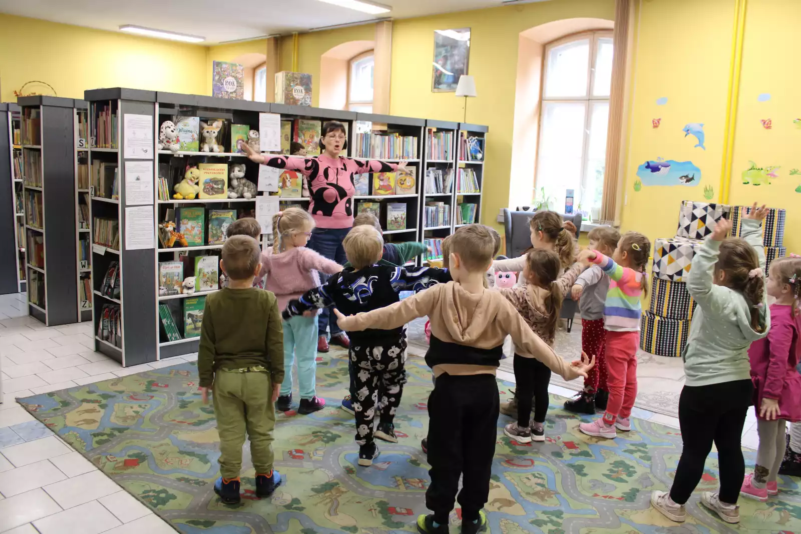 Dzieci stoją na dywanie i powtarzają ruchy wykonywane przez panią bibliotekarkę. 