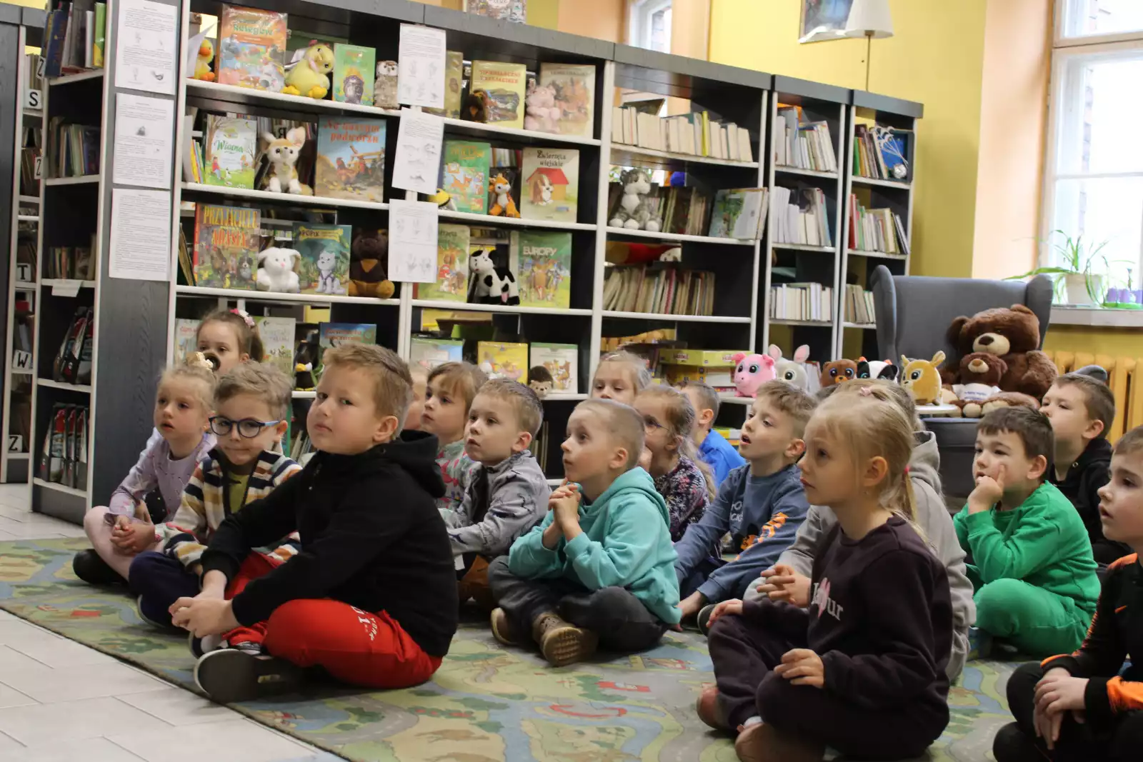 Dzieci siedzą na dywanie i słuchają pani bibliotekarki. 