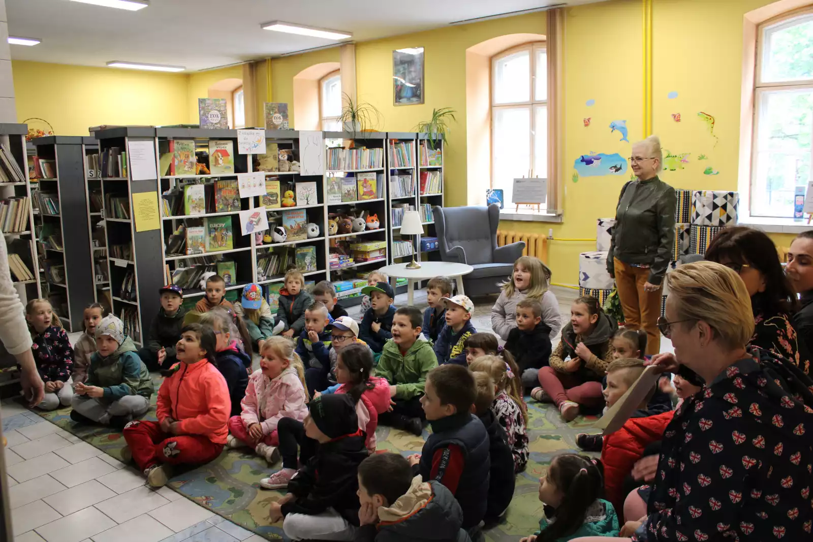 Dzieci wraz z paniami nauczycielkami siedzą w bibliotece dziecięcej. 