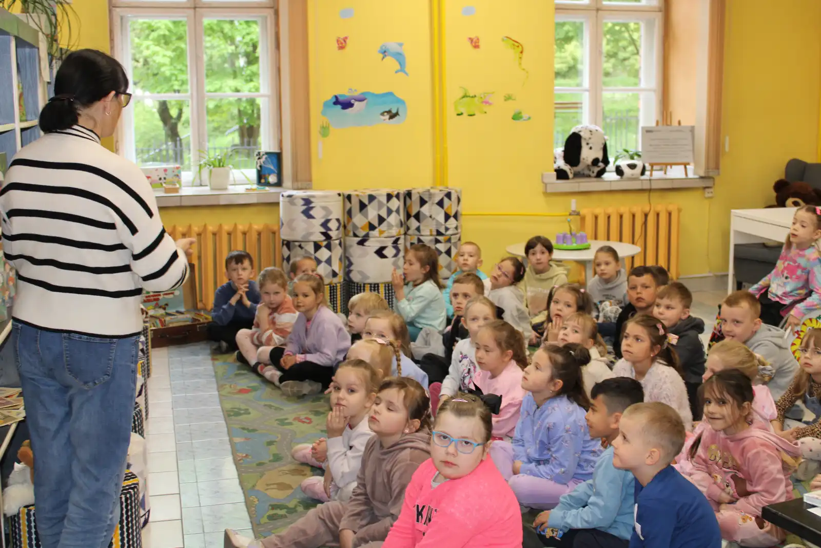 Dzieci siedzą na dywanie i słuchają pani bibliotekarki. 