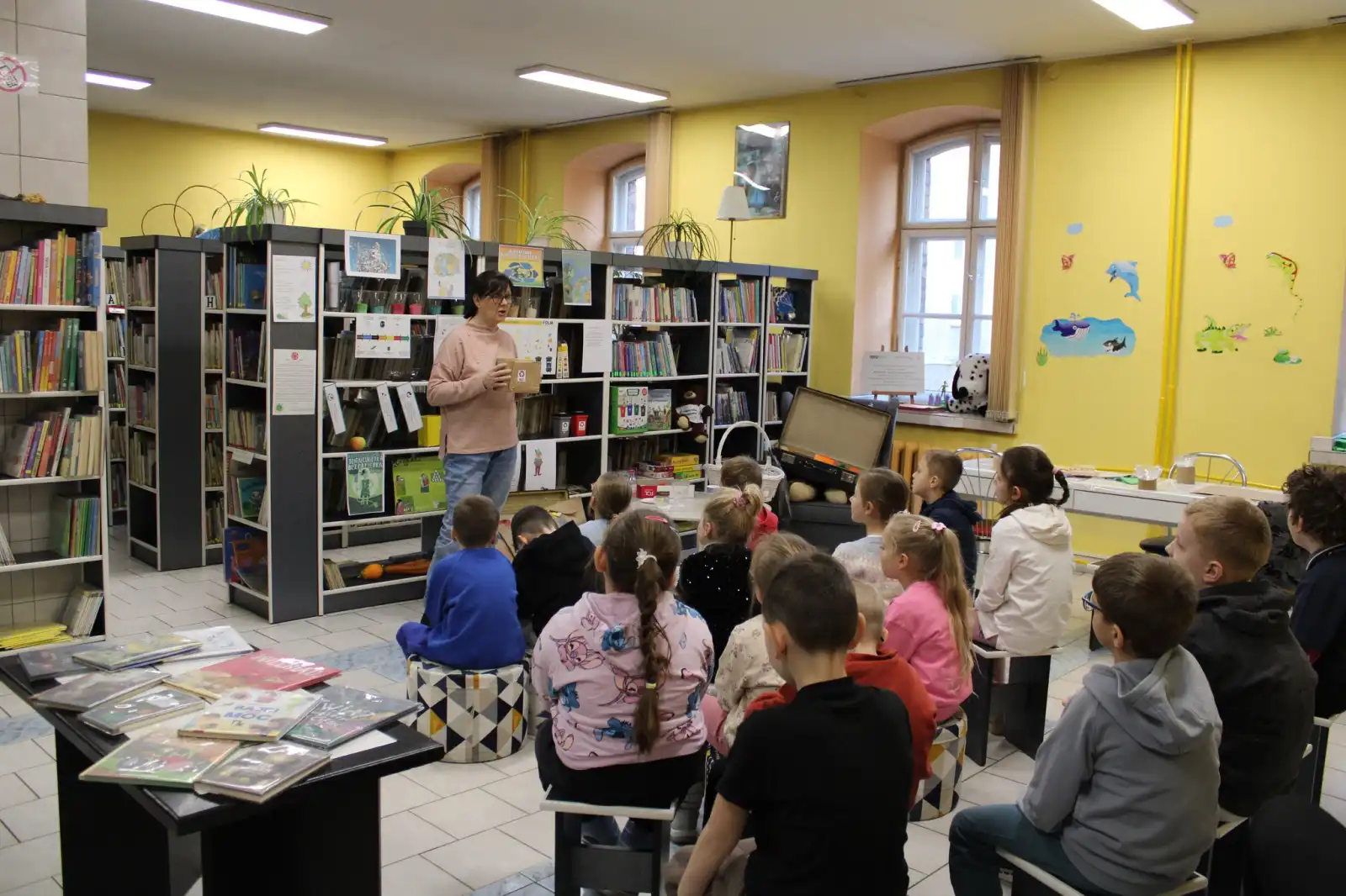 Pani bibliotekarka stoi przed dziećmi, które siedzą na taboretach w Oddziale dla Dzieci.