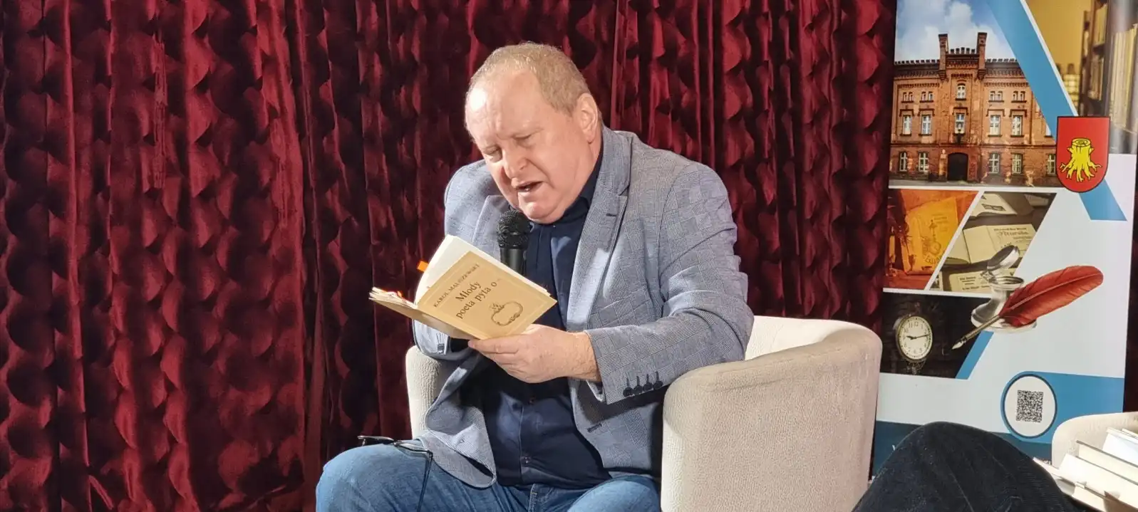 Karol Maliszewski siedzi w fotelu na scenie, czytając swój wiersz.