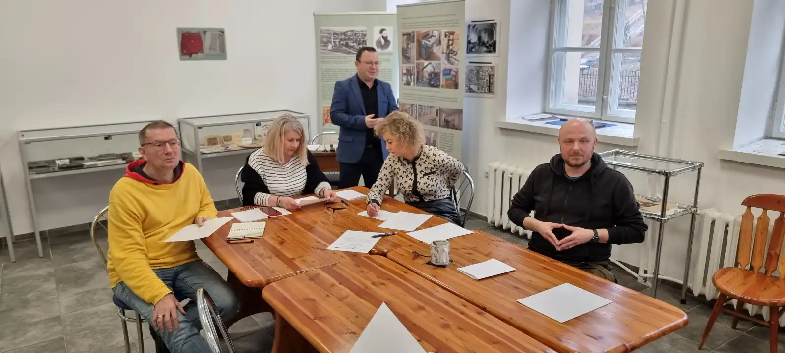 Bibliotekarze siedzą przy stole w trakcie szkolenia. Na drugim planie stoi prowadzący.