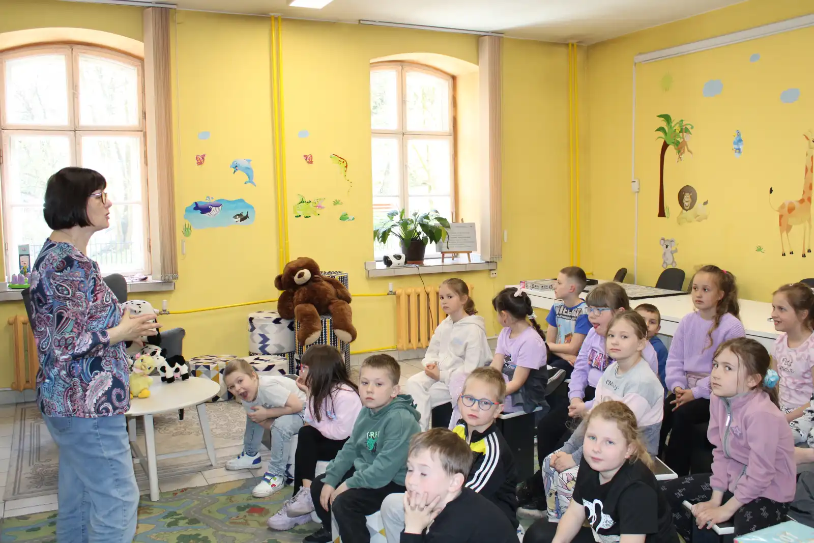 Dzieci siedzą na pufach w Oddziale dla Dzieci. Przed nimi stoi bibliotekarka.